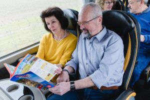 Man und Frau im First-Class-Bus im Pullmann-Doppelsessel sitzend und einen Grimm Reisekatalog lesend