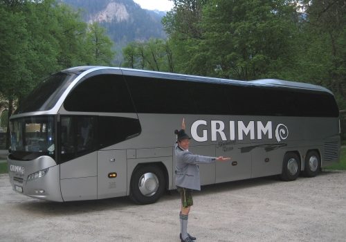 Premium Bus mit GRIMM-Reisen Aufschrift auf einem Parkplatz in den bewaldeten Bergen mit Mann in traditioneller Südtiroler Lederhosentracht