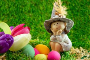 kleine Figur mit Hut sitzt auf Gras mit bunten Ostereiern und Tulpen