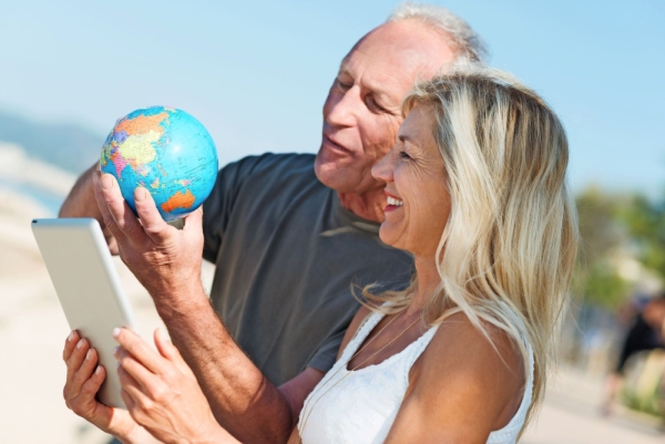 Mann und Frau stehen am Strand, Frau hält Tablet, Mann hält Mini Globus in der Hand, beide schauen auf den Globus